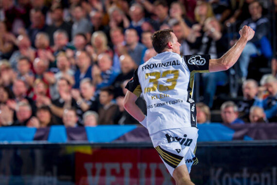 Ein Handballspieler, der ein weißes Trikot mit der Nummer 23 und dem Namen Klein trägt, jubelt mit erhobener Faust vor einer jubelnden Menge während eines Spiels.