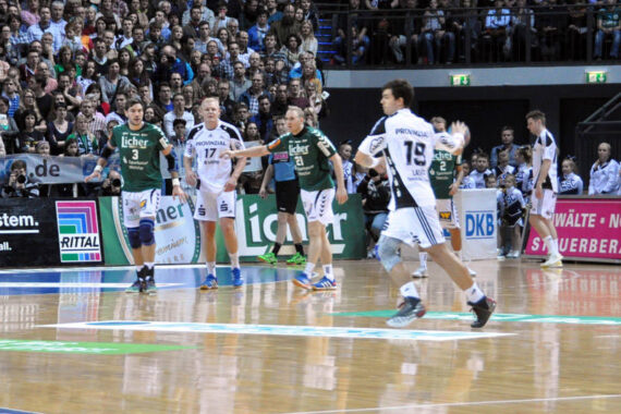 Ein Handballspiel in einer überfüllten Halle; die Spieler in grün-weißen Trikots konzentrieren sich auf den Ball, während die Zuschauer auf der Tribüne aufmerksam zusehen.