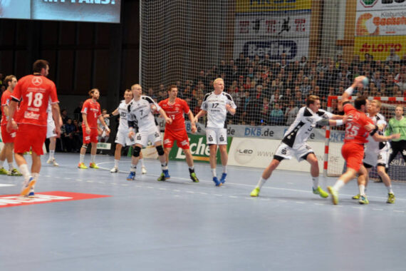 Eine Gruppe männlicher Handballspieler in roten und weißen Trikots tritt in einer Halle gegeneinander an; ein rot gekleideter Spieler ist im Begriff, den Ball zu werfen, während andere ihn verteidigen, während die Zuschauer von der Tribüne aus zusehen.