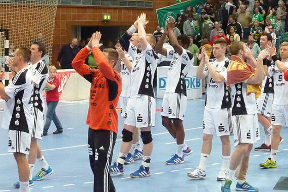 Eine Männer-Handballmannschaft in weißen und schwarzen Trikots klatscht und winkt der Menge auf einem Hallenplatz zu, einige Spieler tragen orangefarbene Trikots. Im Hintergrund sind Zuschauer zu sehen.