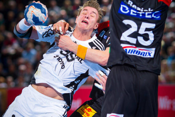 Zwei männliche Handballspieler stoßen während eines Spiels heftig zusammen; der Spieler in Weiß sieht schmerzhaft aus, als er vom Arm des Gegners in Schwarz, der die Trikotnummer 25 trägt, im Gesicht getroffen wird.