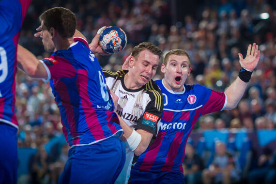 Drei männliche Handballspieler sind während eines Spiels in Aktion. Ein Spieler in einem weiß-schwarzen Trikot hält den Ball, während zwei Spieler in blauen und roten Trikots ihn verteidigen. Im Hintergrund ist eine unscharfe Menschenmenge zu sehen.