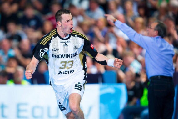Ein männlicher Handballspieler in einem schwarz-weißen Trikot mit der Nummer 33 jubelt leidenschaftlich auf dem Spielfeld, während ein Trainer oder Offizieller im Hintergrund zeigt und schreit. Die Menge hinter ihnen ist unscharf.