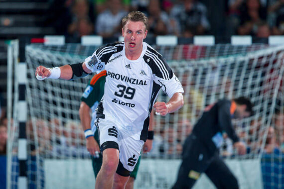 Ein männlicher Handballspieler, der ein weißes Trikot mit der Nummer 39 trägt, läuft während eines Spiels auf einem Hallenplatz, mit einem Tor und unscharfen Spielern im Hintergrund.