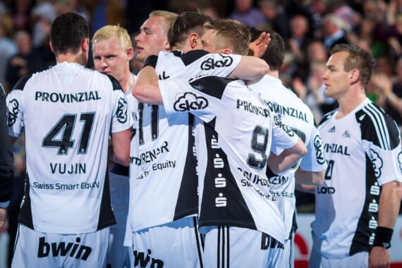 Eine Gruppe männlicher Handballspieler in weißen und schwarzen Trikots umarmt und feiert auf dem Spielfeld, mit dem Rücken zur Kamera, vor einer verschwommenen Menge.