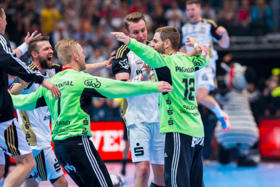 Eine Gruppe von männlichen Handballspielern in Trikots feiert ausgelassen auf einem Hallenplatz, wobei einige Spieler grüne und andere weiße Trikots tragen. Im unscharfen Hintergrund ist eine jubelnde Menge zu sehen.
