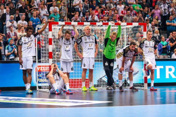 Eine Herren-Handballmannschaft in weißen Trikots steht und sitzt vor dem Tor und sieht nach einem Spiel enttäuscht oder erschöpft aus. Eine Gruppe von Zuschauern schaut von den Tribünen hinter ihnen zu.