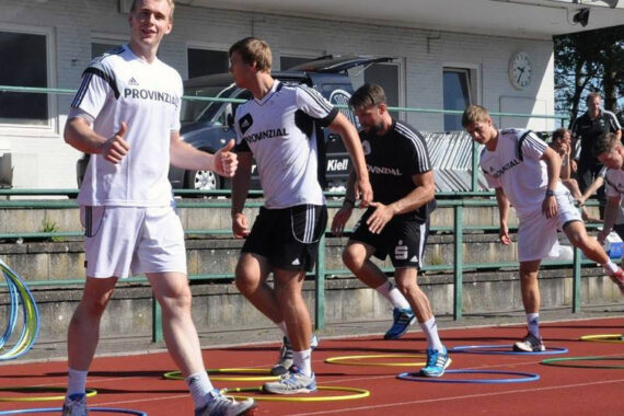 Vier Männer in Sportkleidung machen Beweglichkeitsübungen auf einer Bahn, indem sie durch bunte Reifen auf dem Boden springen. Der Mann im Vordergrund lächelt und winkt in die Kamera. Im Hintergrund sind ein Gebäude und ein Lieferwagen zu sehen.