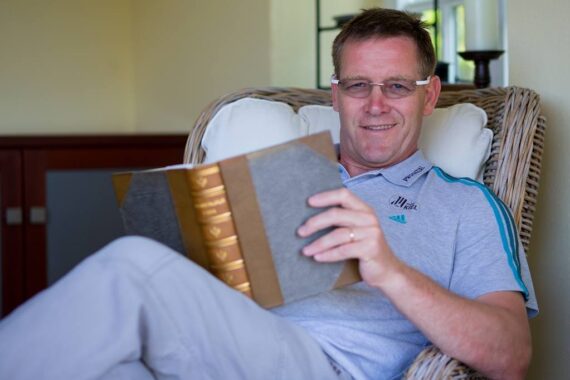 Ein Mann mit Brille und grauem Polohemd sitzt in einem Korbstuhl, lächelt und hält ein aufgeschlagenes Buch in seinem Schoß.