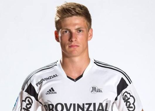 Ein junger Mann mit kurzen blonden Haaren, der ein weiß-schwarzes Sporttrikot mit der Aufschrift Provinzial und verschiedenen Logos trägt, steht vor einem schlichten weißen Hintergrund.