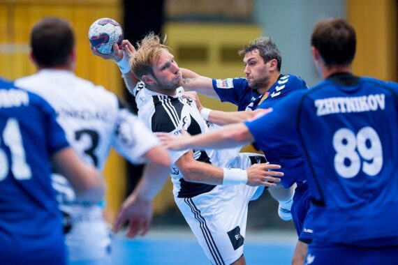 Zwei Handballspieler, einer in Weiß und einer in Blau, stoßen in der Luft zusammen, als sie um den Ball kämpfen. Zwei weitere Spieler in Blau, mit den sichtbaren Nummern 3 und 89, sind ebenfalls in der Szene und beobachten das Spiel.