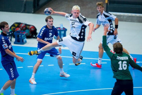 Ein Handballspieler in einem weißen Trikot springt in die Luft, um den Ball zu schießen, während Spieler in blauen Trikots und ein Torwart in Grün versuchen, ihn auf einem Hallenplatz zu blockieren.
