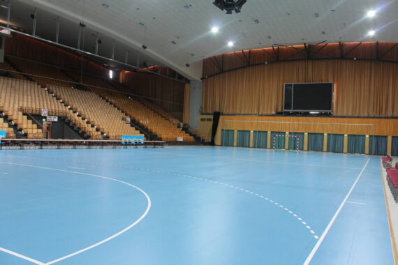 Eine Sporthalle mit einem blauen Spielfeld, leeren Sitzreihen, einer großen Anzeigetafel an der Wand und heller Deckenbeleuchtung. Es sind keine Menschen anwesend.