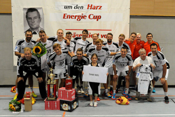 Ein Herrensportteam in schwarz-weißen Uniformen posiert in einer Halle mit Trophäen, Blumen und Geschenkboxen. Dahinter ein Banner mit dem Energie Cup und ein schwarz-weißes Porträt. Eine Person hält ein Schild des THW Kiel in der Hand.