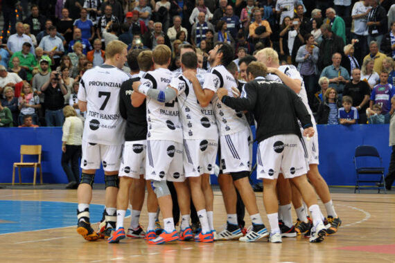 Eine Herren-Handballmannschaft in weißen Trikots kauert in einer Halle zusammen und feiert, umgeben von einer jubelnden Menge auf der Tribüne.