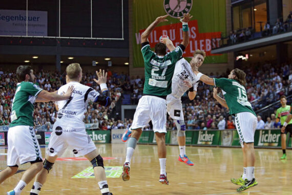 Ein Handballspieler in einem weißen Trikot springt, um den Ball zu schießen, während drei Gegner in Grün versuchen, ihn zu blockieren; im Hintergrund sind ein Schiedsrichter und eine Menschenmenge zu sehen.