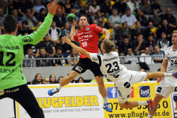 Ein Handballspieler in einem roten Trikot springt, um den Ball zu werfen, während ein Spieler in einem weißen Trikot versucht, ihn in der Luft zu blockieren. Der Torwart und ein weiterer Spieler schauen zu, im Hintergrund sind Zuschauer zu sehen.
