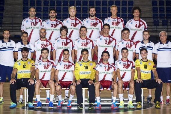 Eine Herren-Handballmannschaft stellt sich für ein Gruppenfoto in einer Sporthalle auf. Die Spieler tragen weiße und rote Trikots, wobei zwei Spieler in gelben Trikots vorne sitzen. Trainer und Betreuer stehen auf beiden Seiten.