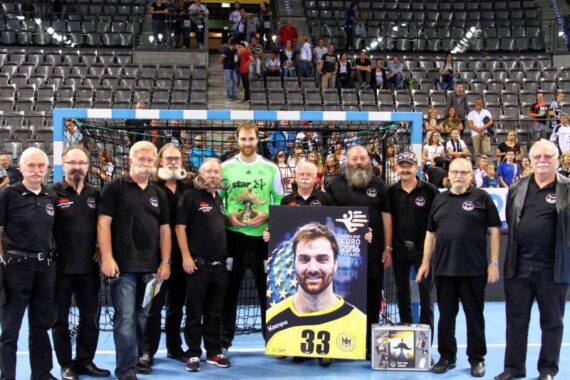 Eine Gruppe von Männern in schwarzen Hemden posiert mit einem Handballspieler in Grün vor einem Netz, der ein großes Foto und Erinnerungsstücke hält. Im Hintergrund sind einige Zuschauer zu sehen, die in einer Arena sitzen.