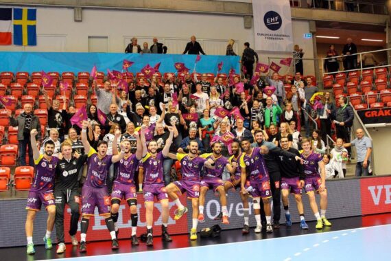 Eine Handballmannschaft in lilafarbenen Trikots posiert freudig vor jubelnden, Fahnen schwenkenden Fans in einer Sporthalle mit roten Sitzen und Bannern, darunter das Logo der EHF (Europäische Handballföderation) im Hintergrund.