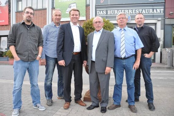 Sechs Männer stehen gemeinsam draußen auf einer gepflasterten Fläche vor einem Gebäude mit einem Schild, auf dem DAS BURGERRESTAURANT steht. Sie posieren für das Foto und sind leger oder formell gekleidet.