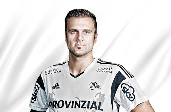 Ein männlicher Sportler, der ein weißes und schwarzes Sporttrikot mit Sponsorenlogos trägt, posiert vor einem weißen Hintergrund und schaut mit ernster Miene geradeaus.