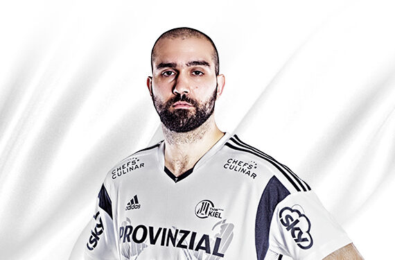 Ein bärtiger Mann in einem weißen Sporttrikot mit schwarzen Streifen und Sponsorenlogos steht vor einem weißen Hintergrund und schaut selbstbewusst in die Kamera.