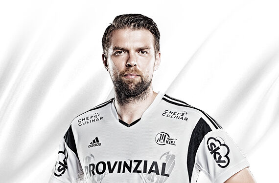 Ein männlicher Handballspieler mit ernster Miene trägt ein weiß-schwarzes THW Kiel Trikot mit verschiedenen Sponsorenlogos und steht vor einem weißen Hintergrund.