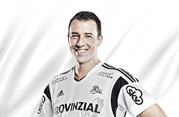 Ein lächelnder männlicher Sportler in einem weißen und schwarzen Sporttrikot mit verschiedenen Sponsorenlogos steht vor einem weißen Hintergrund.