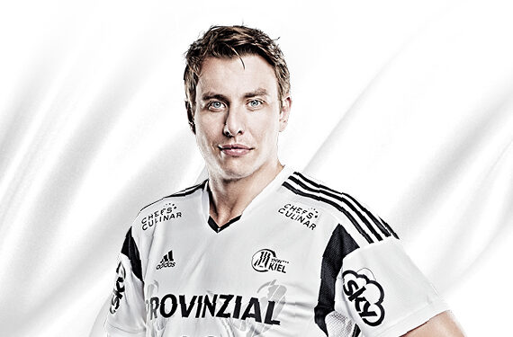 Ein männlicher Sportler in einem weiß-schwarzen Sporttrikot steht vor einem hellen, neutralen Hintergrund und schaut selbstbewusst in die Kamera. Das Trikot ist mit verschiedenen Sponsorenlogos und schwarzen Streifen an den Schultern versehen.