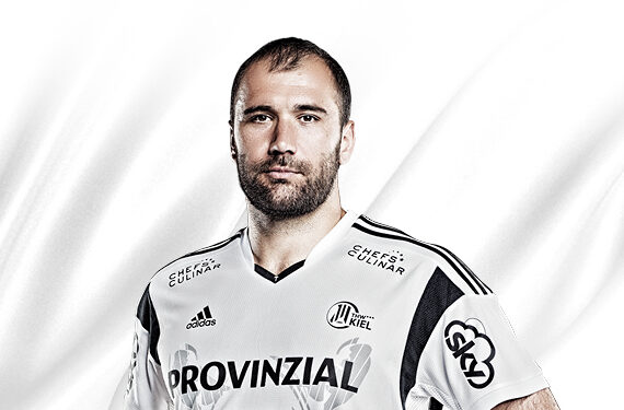 Ein bärtiger männlicher Sportler, der ein weißes Sporttrikot mit schwarzen Streifen und mehreren Sponsorenlogos trägt, steht vor einem schlichten weißen Hintergrund.