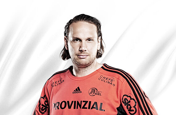 Ein Mann mit schulterlangem Haar, der ein orangefarbenes Sporttrikot mit schwarzen Streifen und Sponsorenlogos trägt, steht vor einem weißen Hintergrund. Das Trikot trägt auf der Vorderseite die Aufschrift PROVINZIAL.