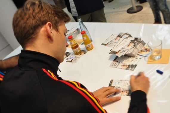 Eine Person in einer schwarzen Jacke mit roten und gelben Streifen sitzt an einem Tisch und gibt Autogramme auf einem Stapel von Fotos. Auf dem weißen Tisch stehen Getränke und ein Stift. Andere Personen stehen in der Nähe.