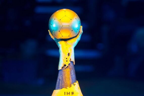 Nahaufnahme einer goldenen Trophäe in Form von zwei Händen, die einen Fußball halten und auf einem Sockel mit den Buchstaben IHF stehen. Der Hintergrund ist unscharf mit dunkelblauen Tönen.