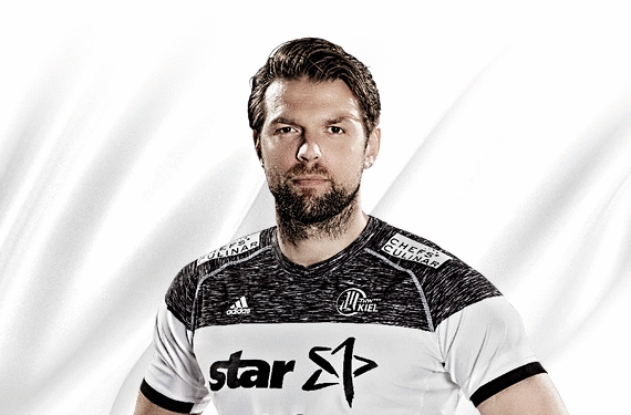 Ein männlicher Handballspieler mit kurzen braunen Haaren und einem Bart posiert in einem weißen und schwarzen Trikot mit Sponsorenlogos vor einem hellen Hintergrund.