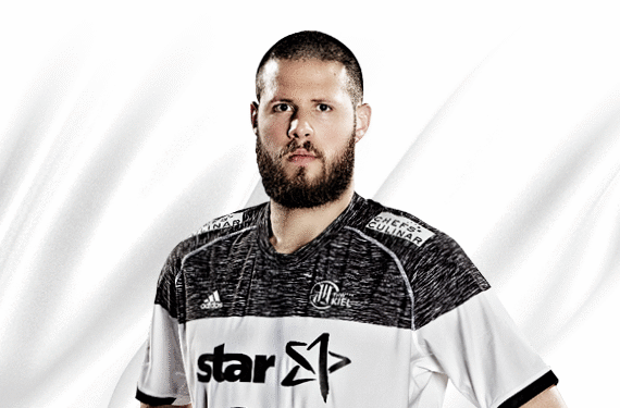 Ein bärtiger Mann in einem schwarz-weißen Sporttrikot steht vor einem weißen Hintergrund und schaut mit neutralem Blick direkt in die Kamera.