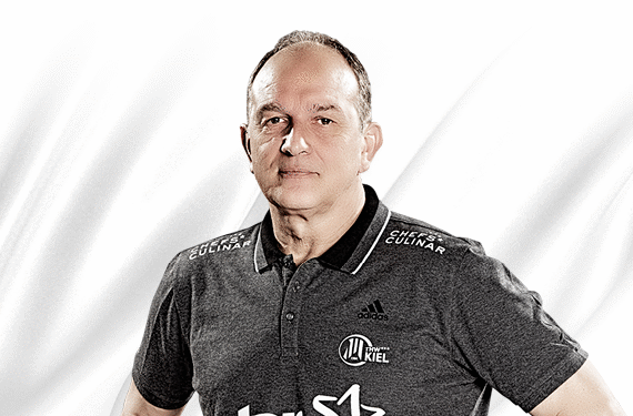 Ein Mann mittleren Alters mit kurzen Haaren und einem dunkelgrauen Poloshirt mit weißen Logos steht vor einem weißen Hintergrund. Auf dem Hemd sind Sport- und Markenlogos abgebildet, darunter Kiel und Adidas.