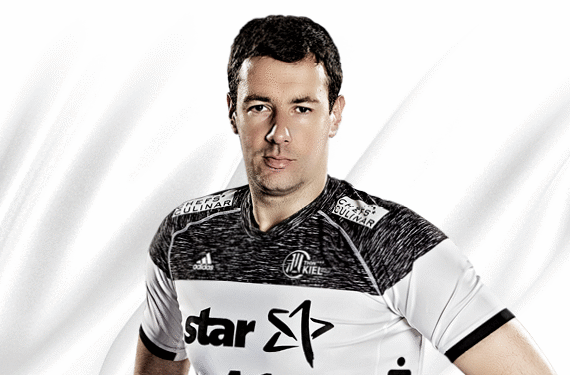 Ein männlicher Sportler, der ein schwarz-weißes Sporttrikot mit dem Wort Star und Logos trägt, steht vor einem weißen Hintergrund und schaut mit neutralem Blick direkt in die Kamera.