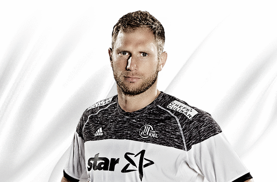 Ein männlicher Sportler, der ein schwarz-weißes Sporttrikot mit Logos trägt, steht vor einem schlichten weißen Hintergrund und schaut direkt in die Kamera.