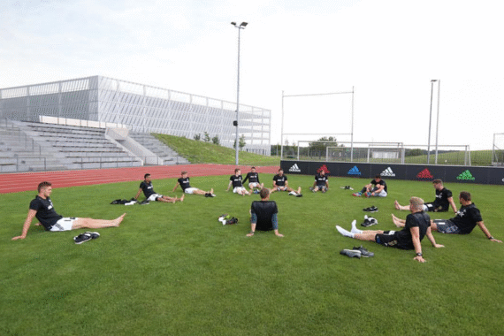 Eine Gruppe von Fußballspielern sitzt im Kreis auf dem Rasen und dehnt sich auf einem Sportplatz. Eine leere Tribüne und ein modernes Gebäude befinden sich im Hintergrund unter einem bewölkten Himmel.