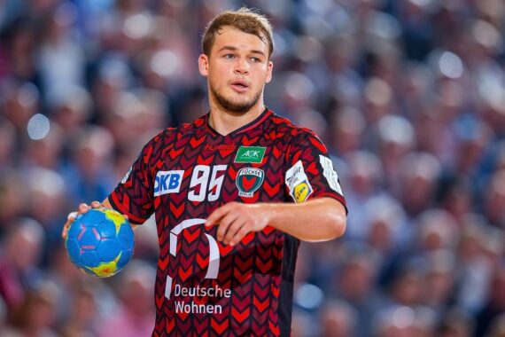 Ein männlicher Handballspieler in einem rot-schwarzen Trikot mit der Nummer 95 hält einen blau-gelben Ball und schaut konzentriert während eines Spiels. Der Hintergrund ist mit einer unscharfen Menge gefüllt.