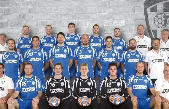 Eine Herren-Handballmannschaft posiert für ein Gruppenfoto in blau-schwarzen Trikots mit Trainern in weißer Kleidung. Sie stehen und sitzen in Reihen vor einer grauen Backsteinwand mit dem TBV-Logo.