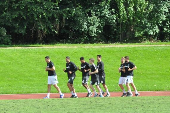 Eine Gruppe von sieben Personen in schwarzen Hemden und Shorts joggt gemeinsam auf einer roten Laufbahn, im Hintergrund grünes Gras und Bäume.