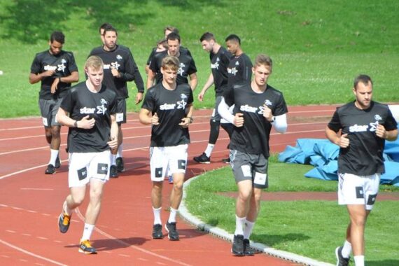 Eine Gruppe junger Männer in schwarzen Hemden und weißen Shorts joggt gemeinsam auf einer roten Laufbahn, auf der im Hintergrund Gras und Sportgeräte zu sehen sind.