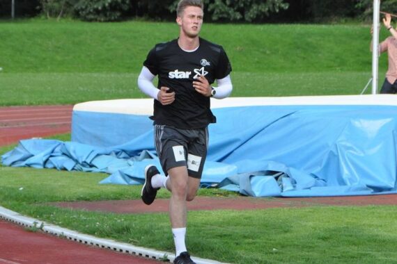 Ein junger Mann in Sportkleidung joggt auf einer roten Laufbahn, neben einer Hochsprungmatte und grünem Gras, mit Bäumen im Hintergrund.