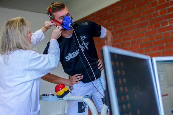 Eine Frau in einem Laborkittel passt eine blaue Atemmaske an einem Mann an, der auf einem Trainingsgerät steht, während im Vordergrund Computermonitore Daten anzeigen. Die Szene spielt sich in einem Raum mit einer Backsteinwand ab.