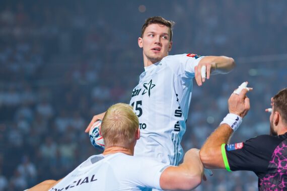 Ein männlicher Handballspieler in einem weißen Trikot springt mit dem Ball und bereitet sich auf den Wurf vor, während zwei gegnerische Spieler in dunklen Trikots versuchen, ihn während eines intensiven Spiels zu blockieren.