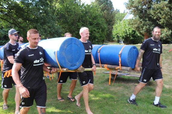 Fünf Männer in passender schwarzer Sportkleidung tragen ein selbstgebautes Floß aus großen blauen Fässern und Holzbrettern über eine Wiese, die an einem sonnigen Tag von Bäumen umgeben ist.
