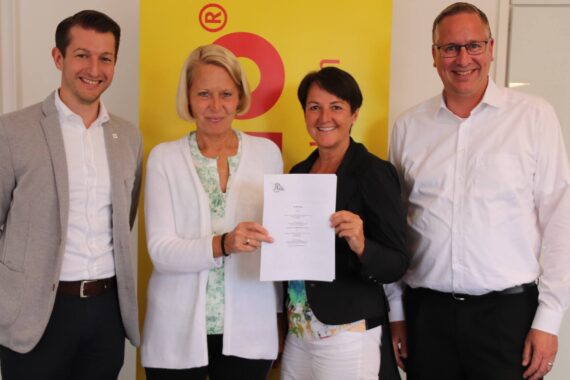 Vier Personen stehen in einem Haus zusammen und lächeln in die Kamera. Die beiden Frauen in der Mitte halten ein Dokument in der Hand. Im Hintergrund ist ein gelbes Schild mit einem roten Logo zu sehen.