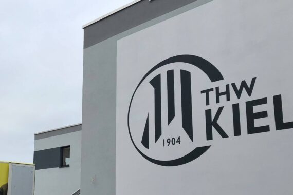 Ein modernes grau-weißes Gebäude mit einem großen Logo des THW Kiel 1904 an der Wand, bei bewölktem Himmel. Links ist ein gelbes Gebäude teilweise sichtbar.